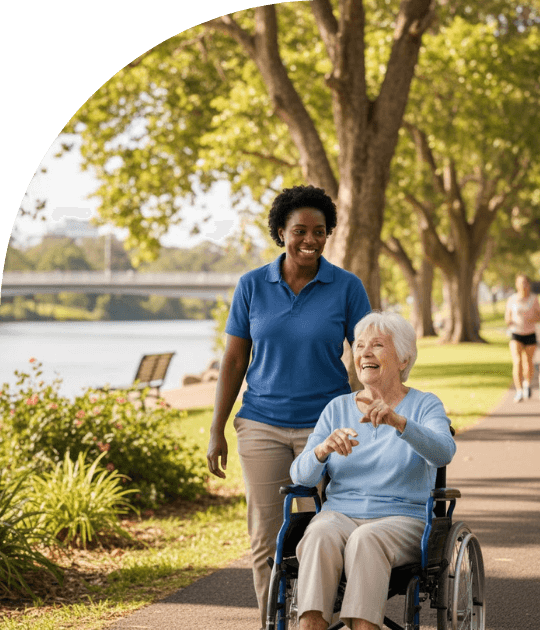 Care worker assists elderly client in a wheelchair on a Bulimba riverside path, promoting community engagement.